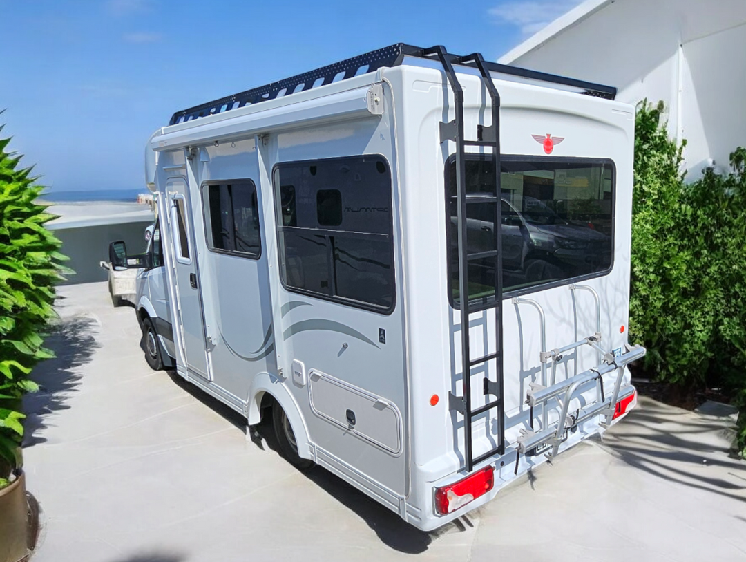 A motorhome with a black powder-coated aluminium roof rack installed on top. The rack has a custom ladder leading up to the roof rack